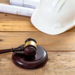 White color engineer safety hardhat, judge gavel and building blueprint plans on wooden table