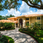 Two-story yellow house with a tiled roof, surrounded by palm trees and gardens, with a curved driveway leading to a front entrance, under a blue sky with clouds.