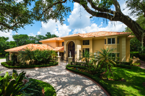 Two-story yellow house with a tiled roof, surrounded by palm trees and gardens, with a curved driveway leading to a front entrance, under a blue sky with clouds.
